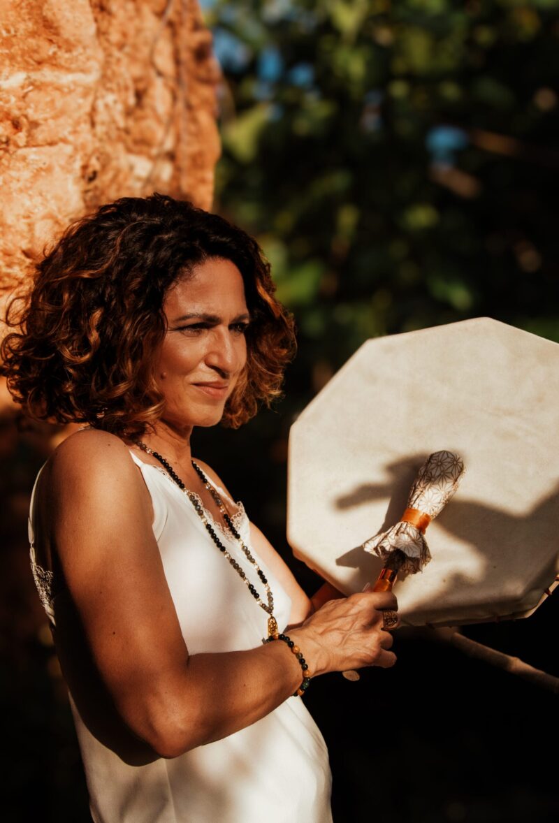 Femme tenant un tambour chamanique, avec un sourire léger et des bijoux spirituels, en arrière-plan naturel.