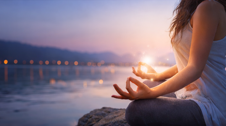 Femme en méditation au bord de l’eau au coucher du soleil, tenant une lumière dans ses mains, représentant l’énergie intérieure, l’alignement et la connexion spirituelle.