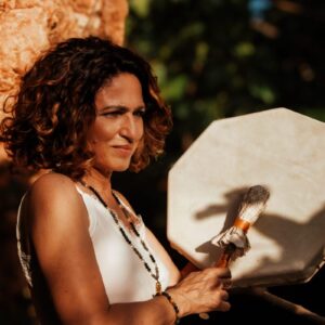 Femme tenant un tambour chamanique, avec un sourire léger et des bijoux spirituels, en arrière-plan naturel.